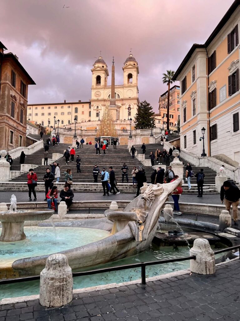 A fountain and the Spanish steps with people on them are illuminated at golden hour, the sky behind the church is pink