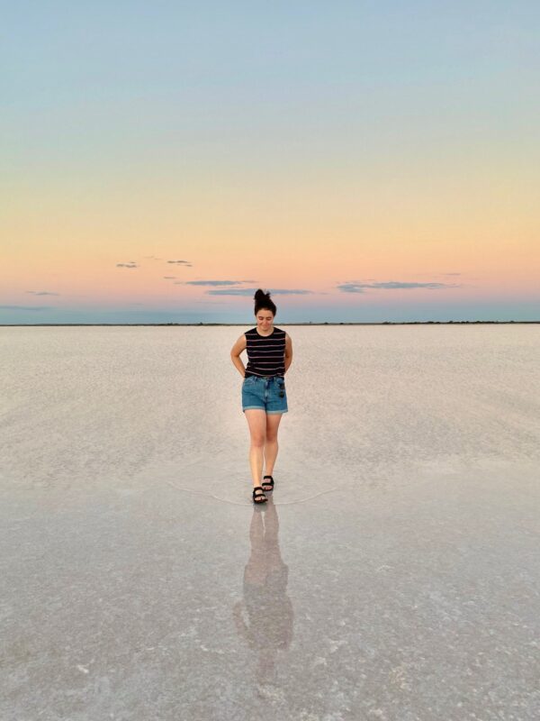 Jenelle walks across a saline lake in Australia at sunset, her reflection shows in the shallow water