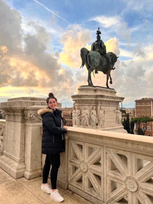 A girl in a puffer jacket stands at the top of the Vittoriano monument in Rome at sunset.