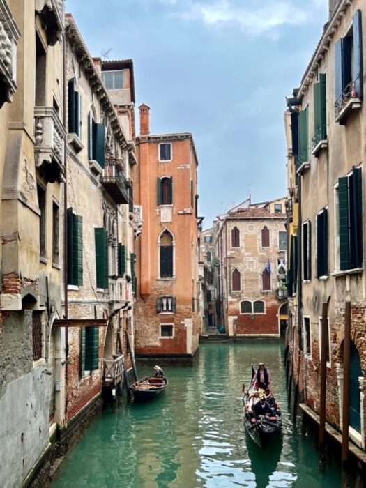 Old buildings with Venetian windows surround a canal filled with green water, a gondolier steers his gondola with passengers.
