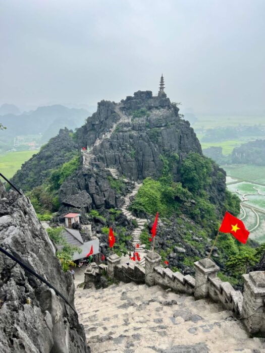 A snaking path of stoney steps leads to a mountain peak in the distance with a pagoda atop it, Vietnamese flags line the path