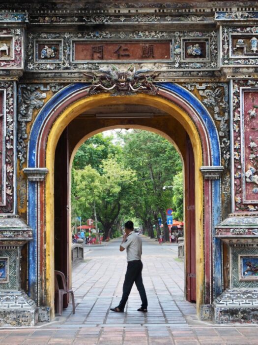 A guard walks through an ancient colourful, decorated gate in the Hue Citadel with a tree lined avenue behind him