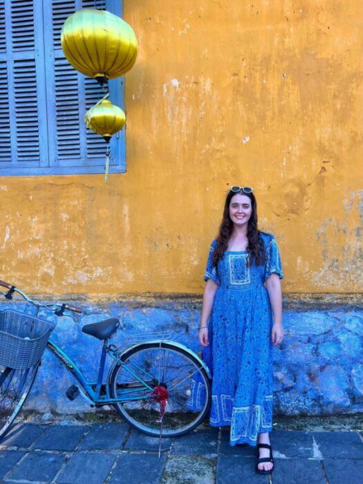 A girl in a blue patterned dress stands against a yellow wall next to a bicycle under a lantern