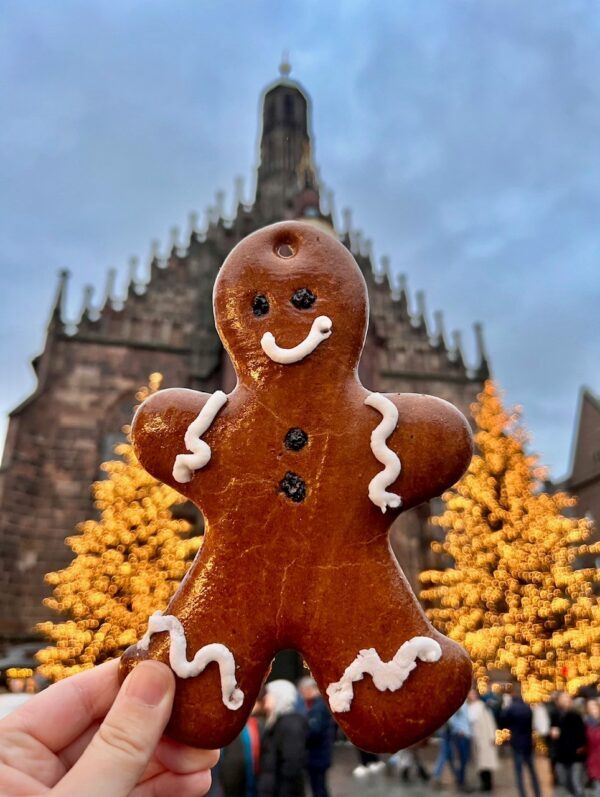 A hand holds up a decorated gingerbread man in front of lit up Christmas trees and a cathedral in Nuremberg