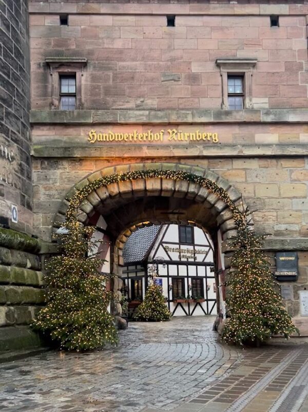 An amber stone archway lined with greenery and fairy lights has golden lettering above it stating Handwerkerhof Nürnberg