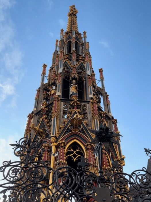 A gilded red and blue fountain stretches up to the sky with iron fretwork in the foreground.