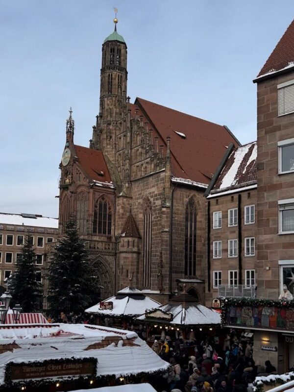 The Nuremberg Christkindlesmarkt tents are covered in snow, behind them the Frauenkirche stands tall with its red roof.