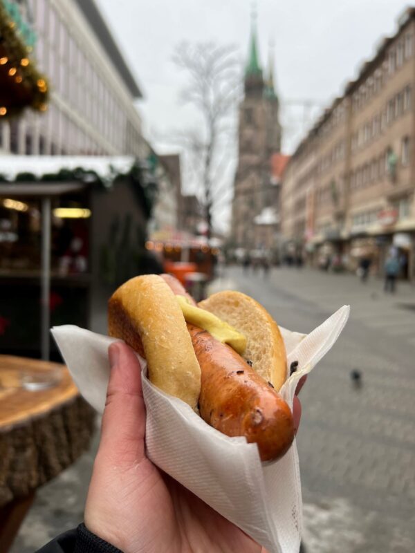 A hand holds a steaming bratwurst with mustard in a bun on the streets of Nuremberg