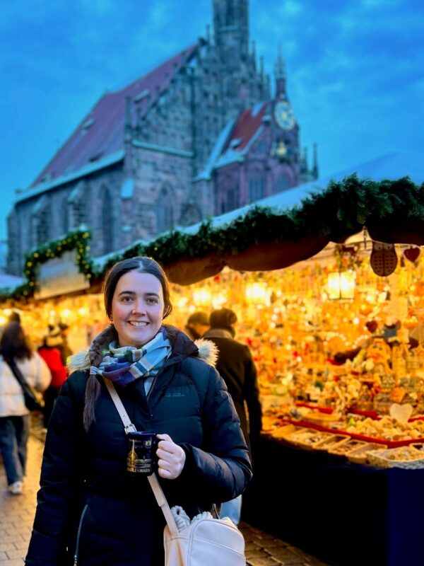 A lady in a puffer jacket holds a mug in front of glowing Christkindlmarkt stalls in Nuremberg, Germany