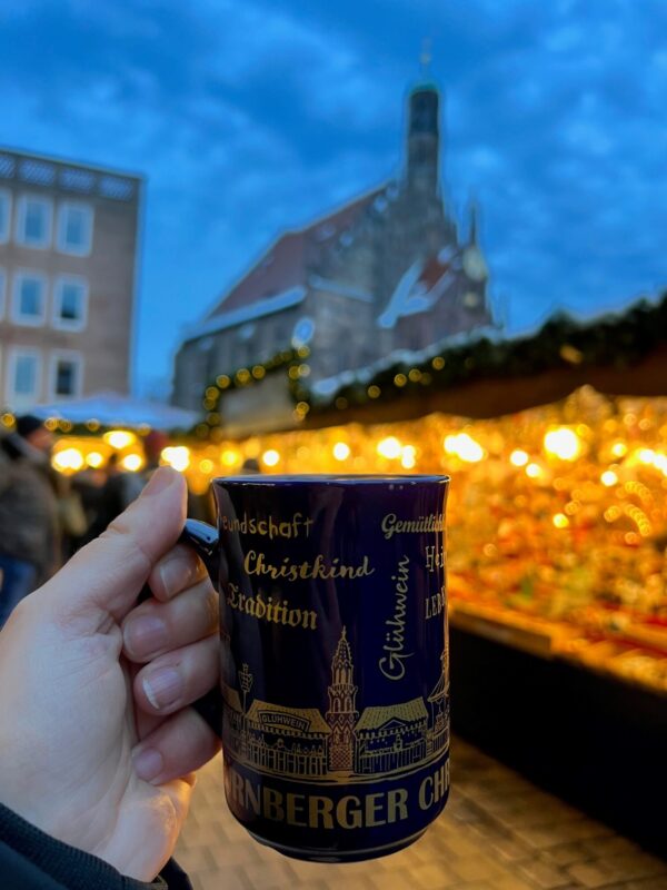 A hand is holding a mug of mulled wine with Christmas market stalls and a church in the background, answering the question is Nuremberg worth visiting.