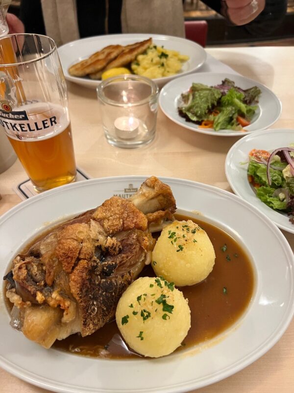 A roasted pork knuckle or Schweinshaxe is on a plate with gravy and two steamed dumplings.