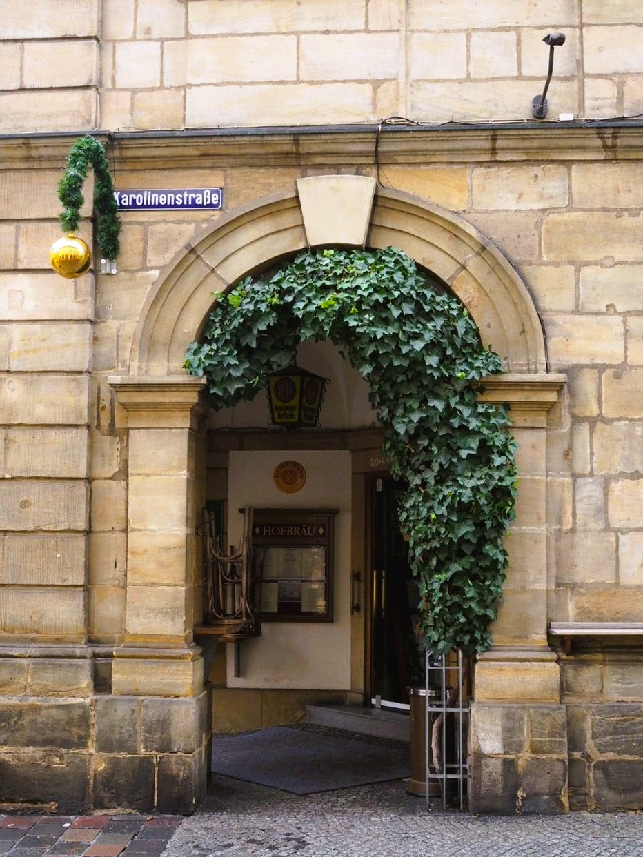 A golden sandstone archway has a deep green ivy plant growing around it and a Christmas decoration hanging above it. A wall sign states Karolinenstrasse