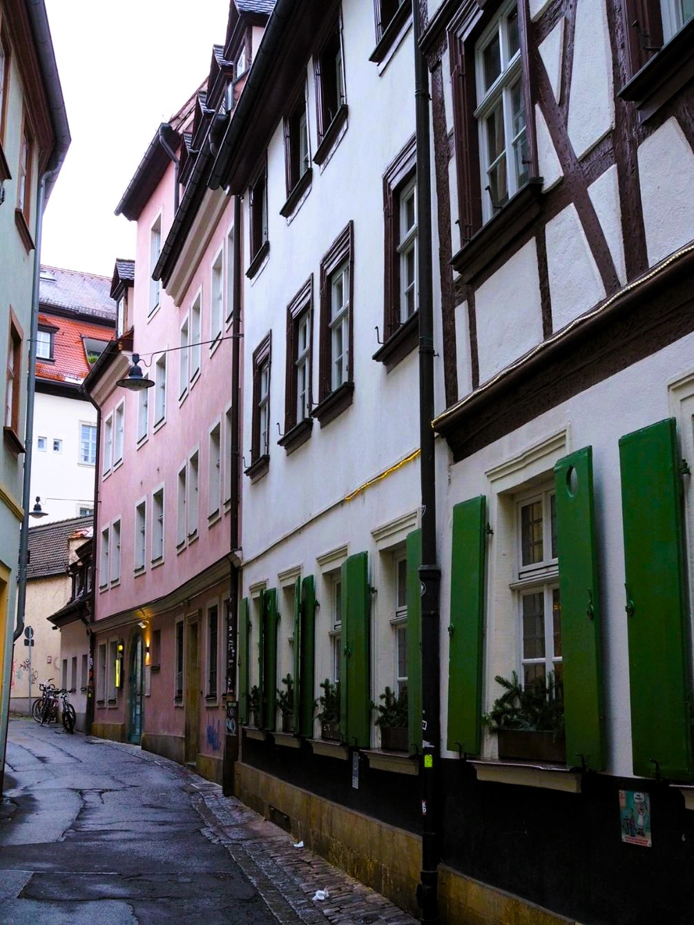 A small alleyway curves away from the camera, a white half-timbered house with green shutters neighbours a pink house with white windowframes in a charming scene.