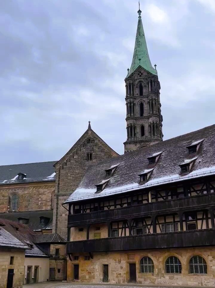 The blue green spire and tan wall of the Bamburg Cathedral ajoin the dark wood and sandstone courtyard of the Alte Hofhaltung, snow dusts the roofs.