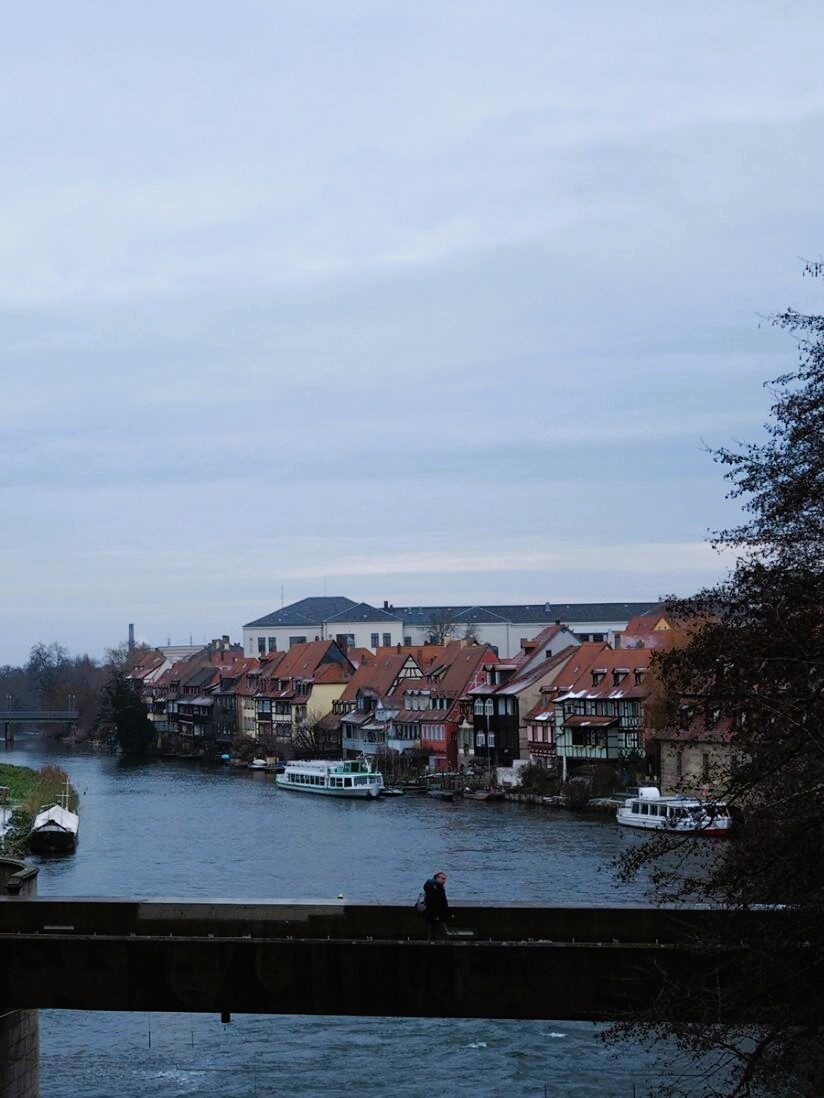 A lone pedestrian walks along a footbridge with a river and multicoloured cottages behind them, small boats are moored in front of the houses.