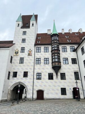 Standing out in a large courtyard filled with cobblestones, a large palace is covered in an diamond pattern of white and grey, topped with copper spires and two coats of arms, beneath the tower, a passageway leads to the road.
