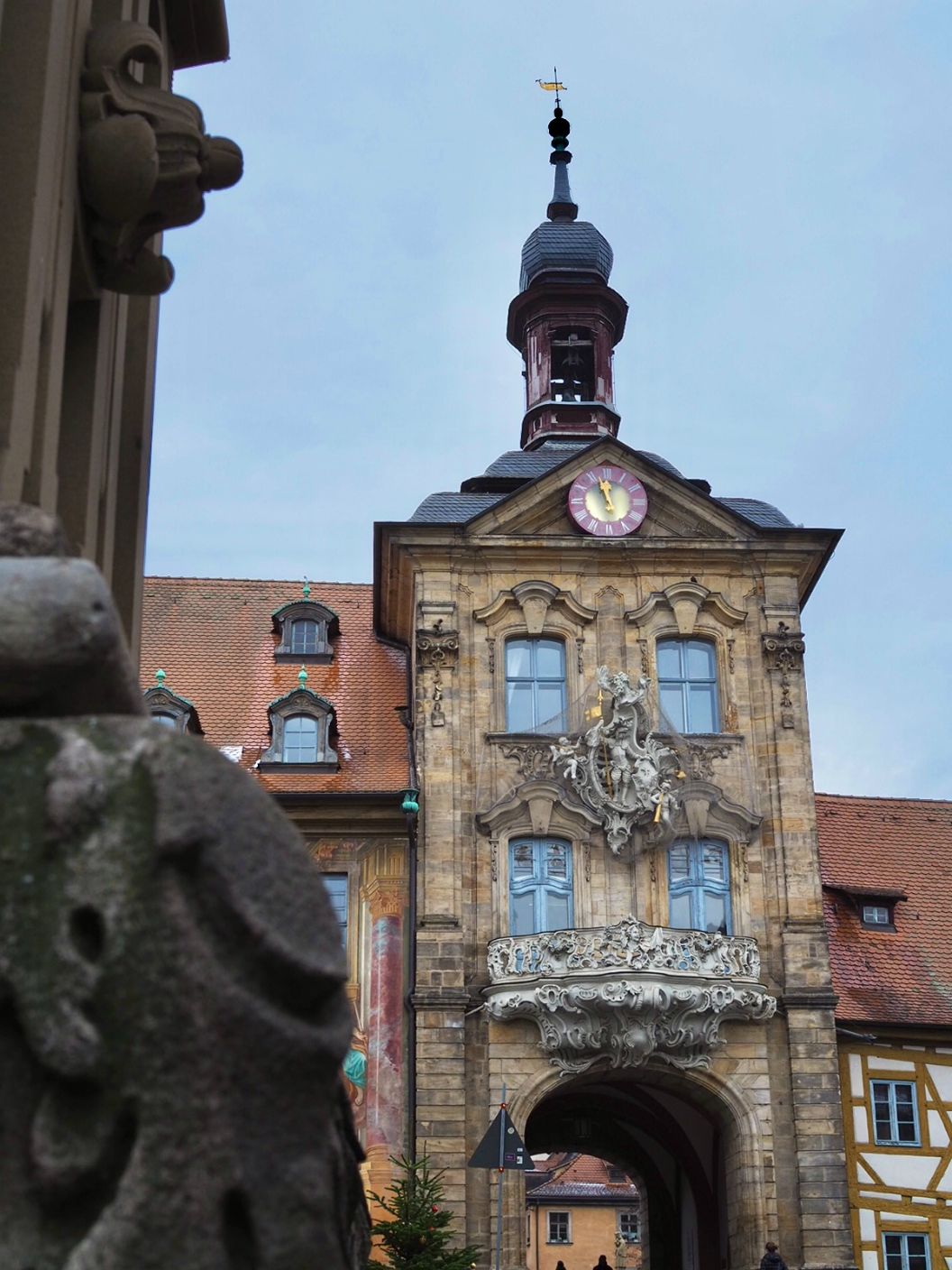 A large Gothic building is shown with a tower and spire atop it, as well as ornate sculptures and a balcony on its facade, a small clock lies above the sculptures.