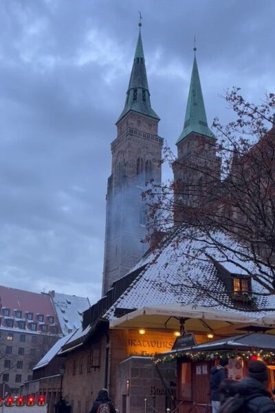 A little hut covered in snow with bright lights and smoke coming out of the chimney serves bratwurst in front of the twin spires of the Sebalduskirche in Nuremberg