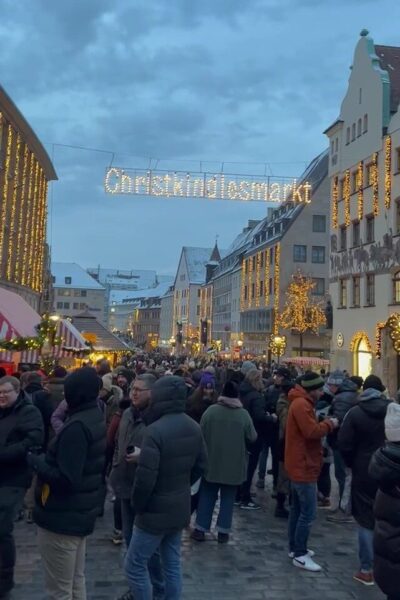 A large crowd of visitors dressed in winter coats stand under a lit up sign stating Christkindlesmarkt.