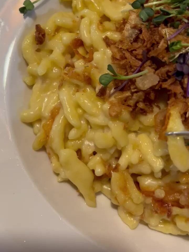 A white plate is shown filled with golden, cheesy noodles, topped with crispy fried onion and microherbs.