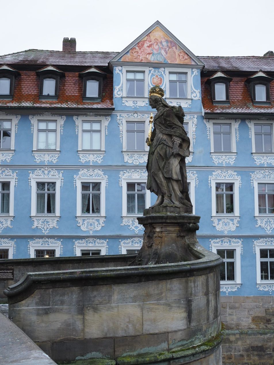 A darkened statue of a woman in flowing robes and a golden crown stands in front of a white and blue filigree decorated town house.