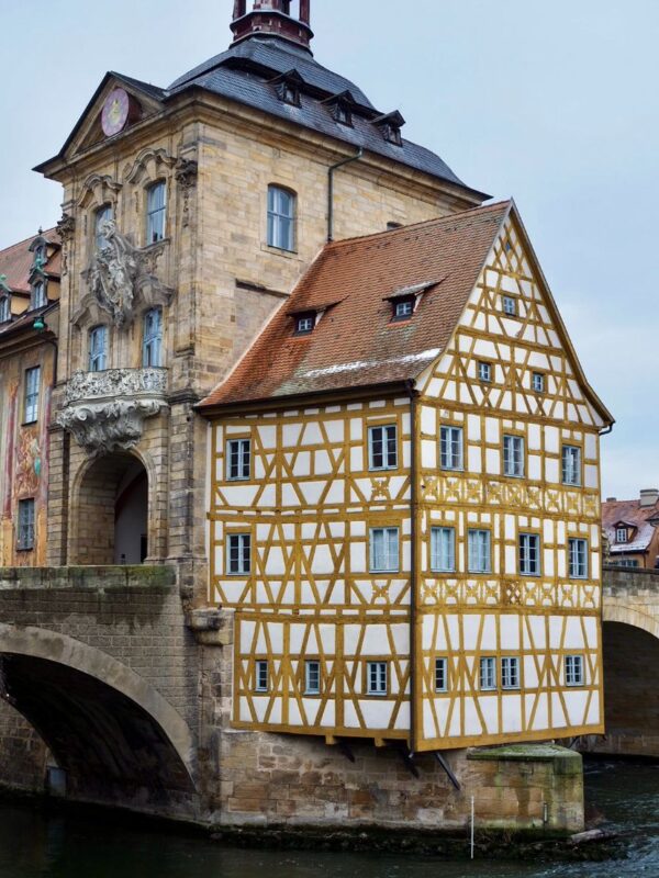 The tan bricks of the Gothic Altes Rathaus in Bamberg is shown with the yellow timbered guard house dangling out over the dark river water.