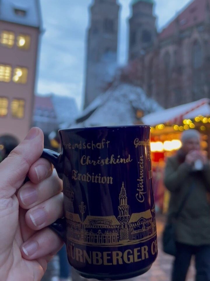 A hand holds a souvenir Gluhwein mug from the Nuremberg Christkindlesmarkt in front of the Sebalduskirche towers.