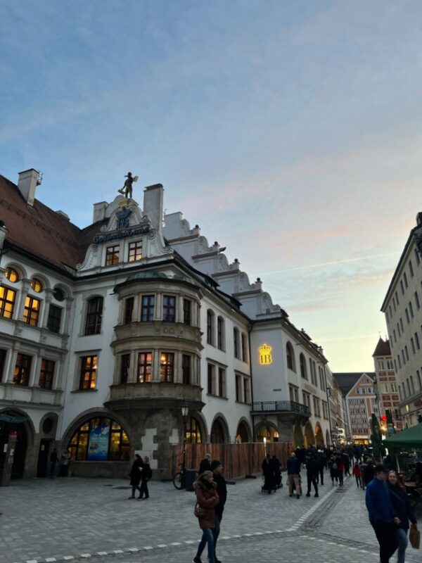 A city square is pictured at twilight, some pink clouds streak a blue sky, within the square, the state owned beer hall has white walls and a distinctive rounded bay window, with a glowing set of initials saying HB, visitors mill below.