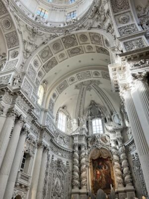 Light plays off the bright white carvings and columns of the Theatinerkirche, flowers and cherubs drape ceilings and columns alike, an altar and organ can just be seen at the bottom of the frame.