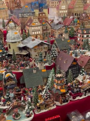 Many snow dusted Christmas porcelain ornaments are displayed on shelves, mostly christmas trees and decorated half timbered houses in statue form.