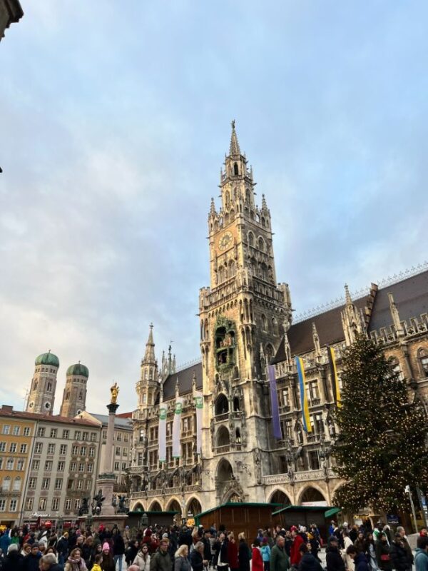 The spires and ornate stonework of the New Town Hall soars into the sky at golden hour, surrounded by visitors, multi-coloured townhouses line the square, complete with a Christmas tree.