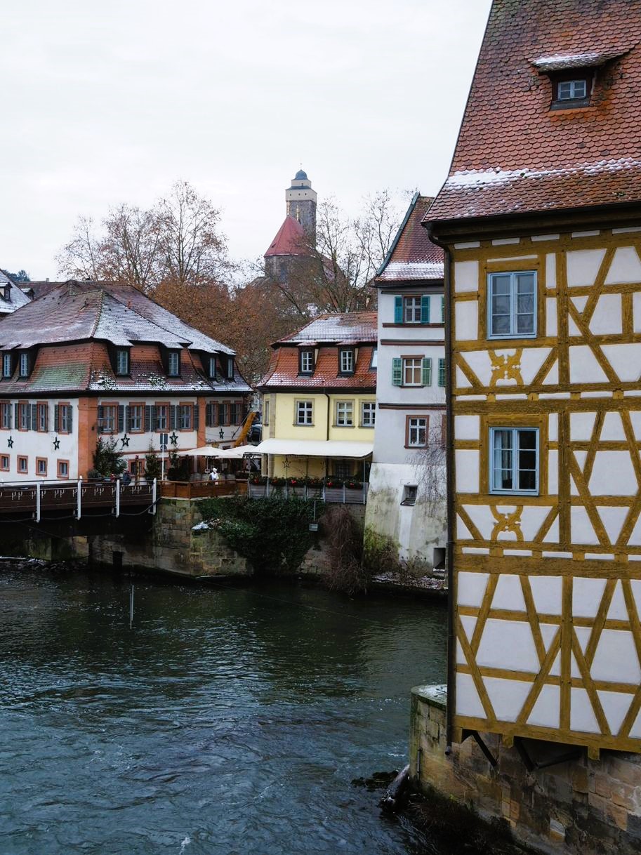 Multiple townhouses are shown fronting the river in Bamberg, all dusted in snow, a church tower looms in the distance.