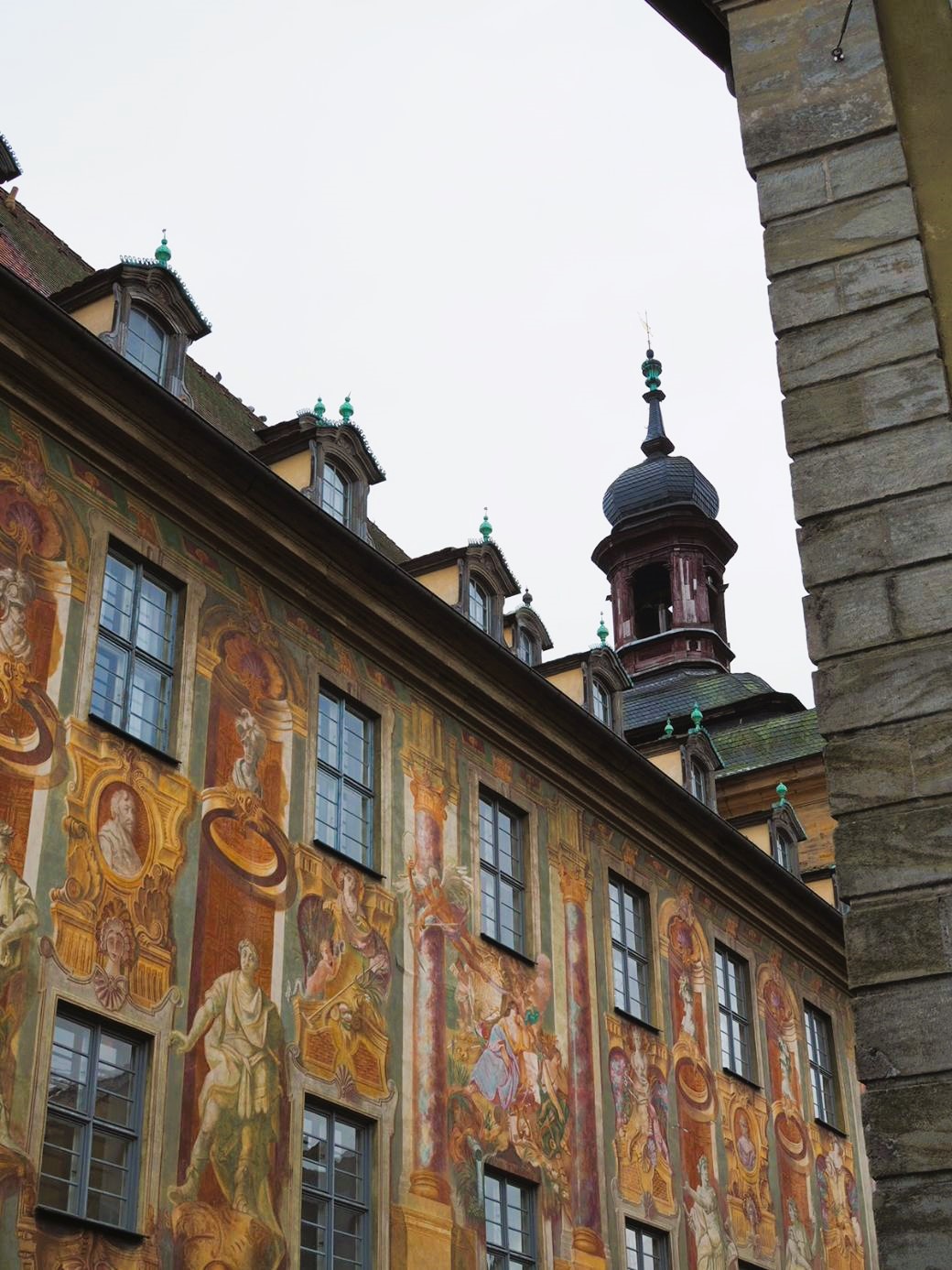 Many multi-coloured murals featuring robed figures and angels are painted onto the walls of the Rathaus in Bamberg, the iron-tipped tower looms over them.