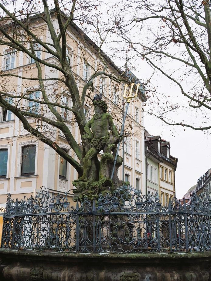 A moss-covered statue of Neptunes stands proud above a fountain, gold tipped trident in his hand, the fountain is surrounded by iron fretwork and a pigeon sits on his head.