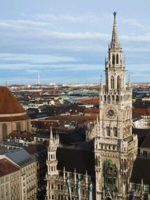 A large, multispired building rises above the rooftops of Munich, a tiny statue on the very top of a small child is copper green, there is also a small clockface on the facade.