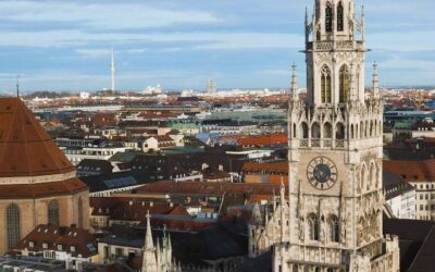 A large, multispired building rises above the rooftops of Munich, a tiny statue on the very top of a small child is copper green, there is also a small clockface on the facade.
