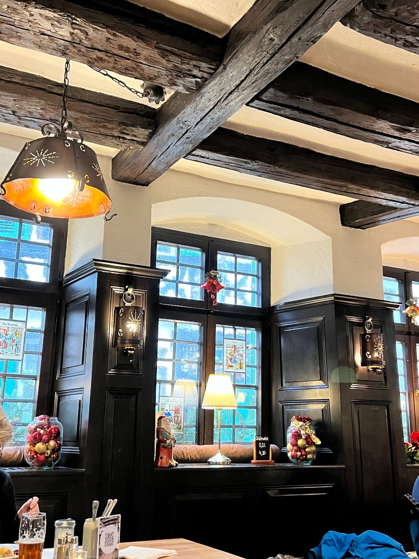 Dark rafters and equally dark wood panelling ring the inside of an ancient dining room of a tavern, lanterns light the cosy scene.