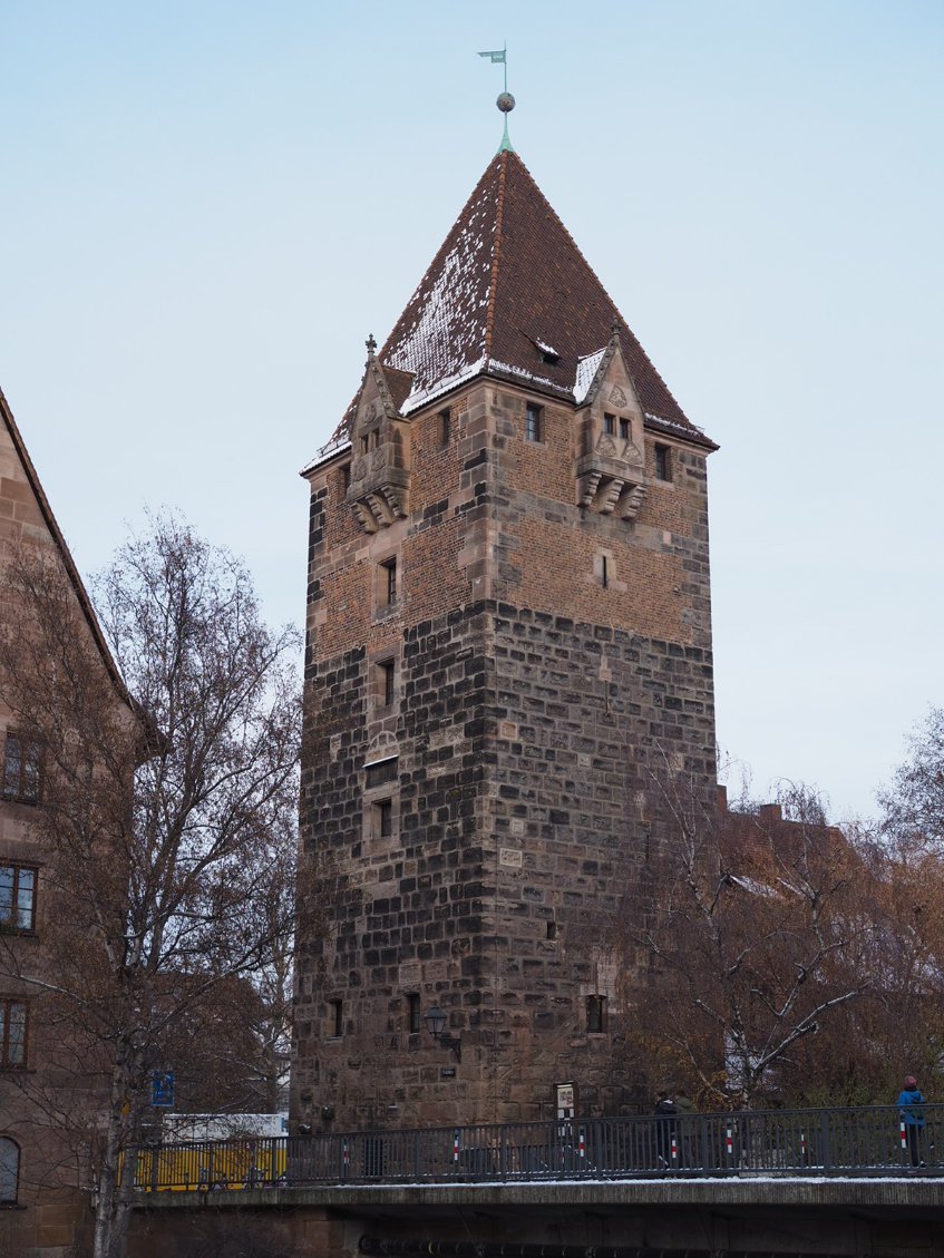 A large rectangular tower with both dark and light coloured stones rises from the old town of Nuremberg, a small flag decorates the spire and snow dusts the roof.