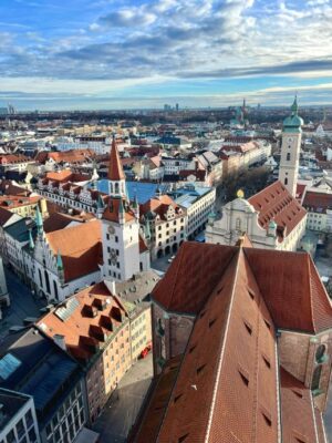 From a bird's eye view, looking down over the red roof of Peterskirche and the copper spires of other old town buildings back to the factories of Munich under a blue morning sky dotted with clouds