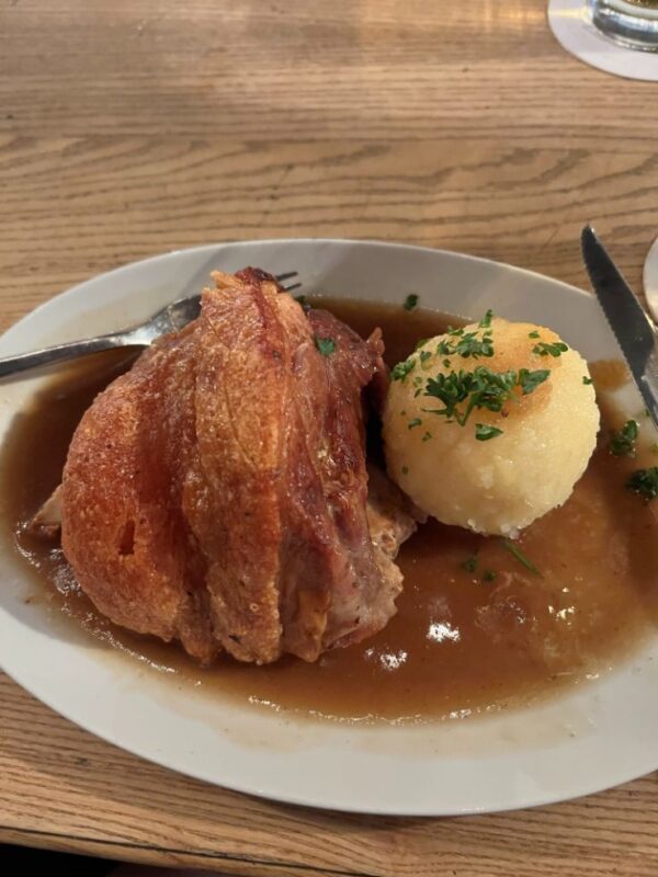 A roasted golden brown pork knuckle with crispy crackling sits on a white plate with a fluffy white dumpling, dusted with parsley and surrounded by gravy, a knife and fork also rest of the plate
