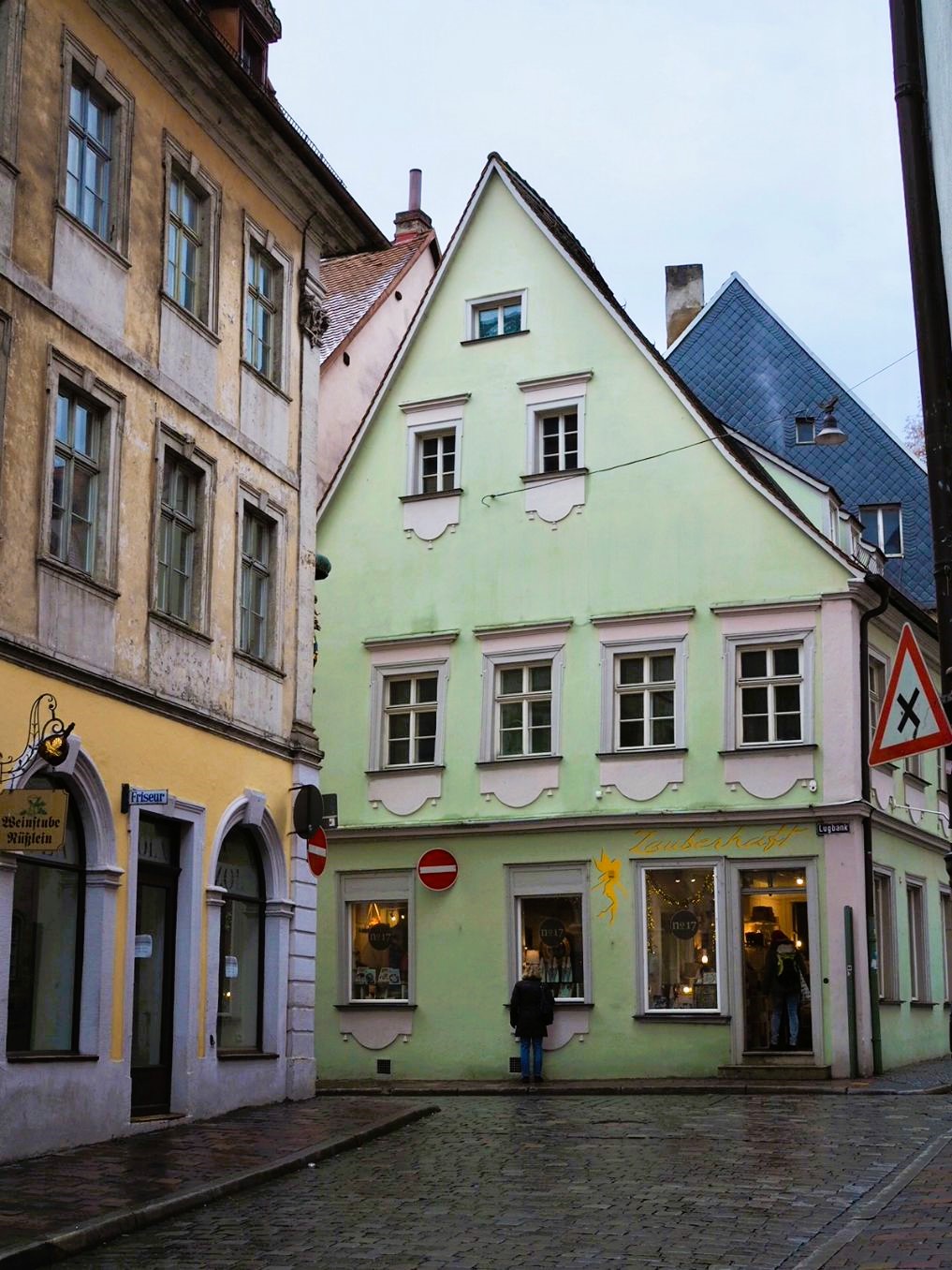 Two multi-storey townhouses are shown in the old town, one painted sunshine yellow and the other a mint green, both house shops on the first floor and have decorated window frames.