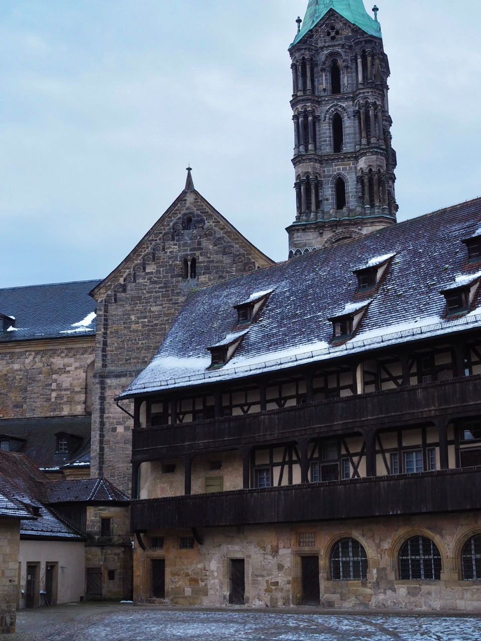 A medieval palace made of sandstone with rows of windows emerging from its dark roof shivers in the shadow of a cathedral tower, the scene is lightly dusted in snow