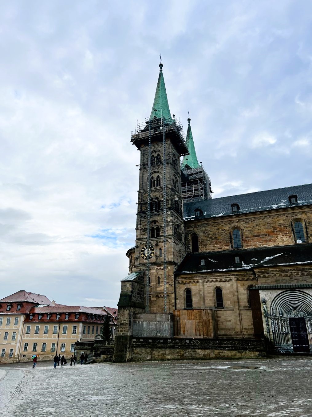 A large medieval cathedral made from sandstone abuts two large towers, topped with blue-green copper spires and scaffolding, seen across a large square.