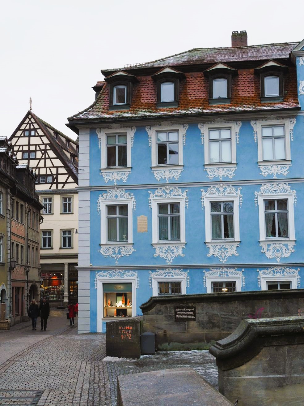 A large house is shown painted duck egg blue with delicate lacework window garnishings, one window is illuminated on the ground floor and glows yellow against the winter gloom.