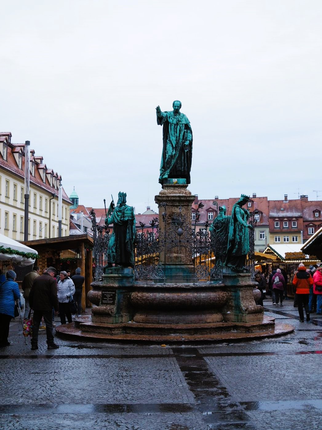 A large fountain is shown topped with five blue green copper statues in various poses, one is higher than the other four with his arm raised. Behind the fountain, crowds enjoy Bamberg's Christmas market in Maximilianplatz.