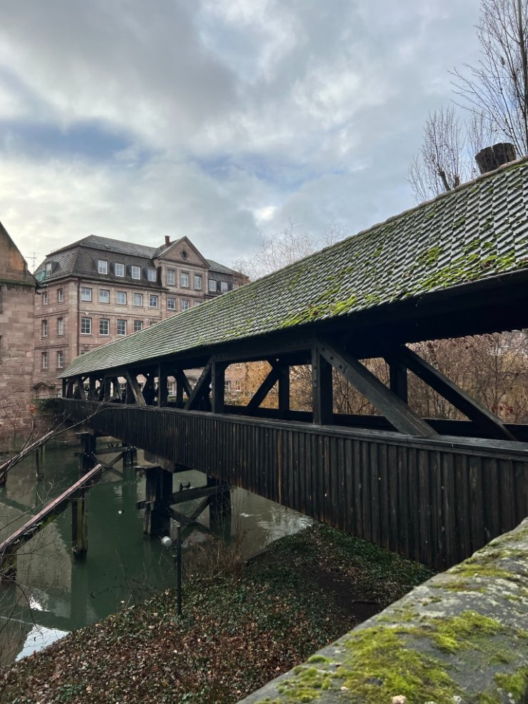 A moss covered bridge with a tiled roof stretches across the Pegnitz River in Nuremberg under a cloudy sky.