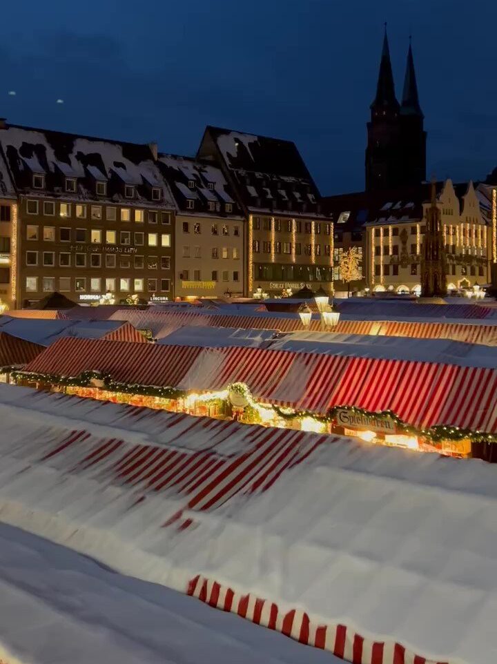 The main square of Nuremberg is lit up at night, filled with red and white striped tents and festive lights on the town houses which surround the stalls. Snow dusts the scene.