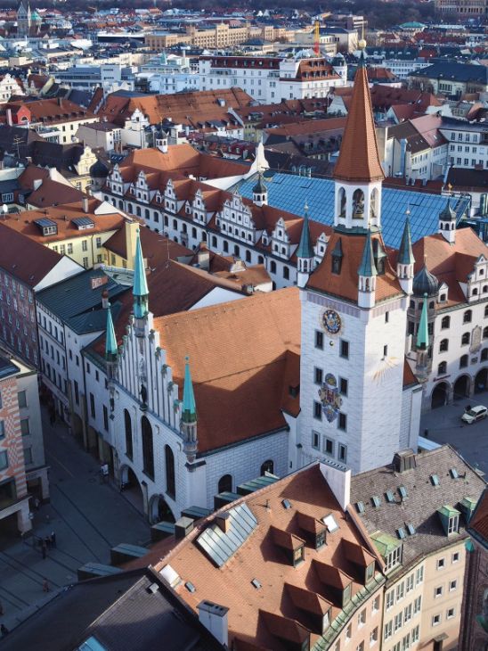 A view over a medieval cityscape taken from above, multiple buildings are shown with quaint red roofs and copper turrets, including the tall Talburgtor tower, decorated with a coat of arms and clock.