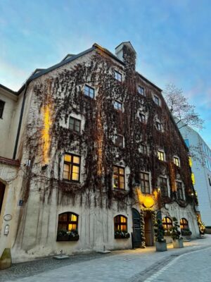A large 5 storied house stands tall against the twilight, lower windows aglow, draped in sleeping vines. See sights like this on the best self-guided walking tour of Munich