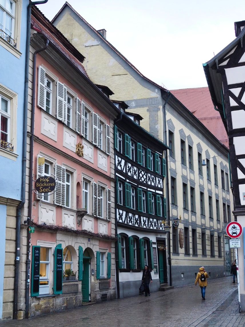 Large townhouses line a cobble stoned street in the centre of the old town, the houses are in lovely pastel shades of pink, blue and yellow, green shutters line the windows. A visitors in a yellow coat strolls through the scene.